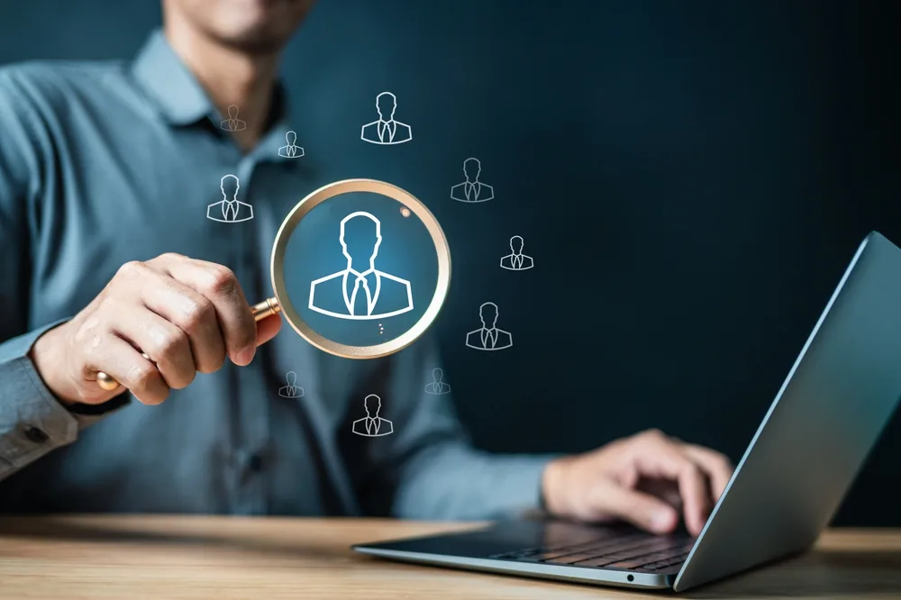 A hand holding a magnifying glass over a candidate icon, above a laptop